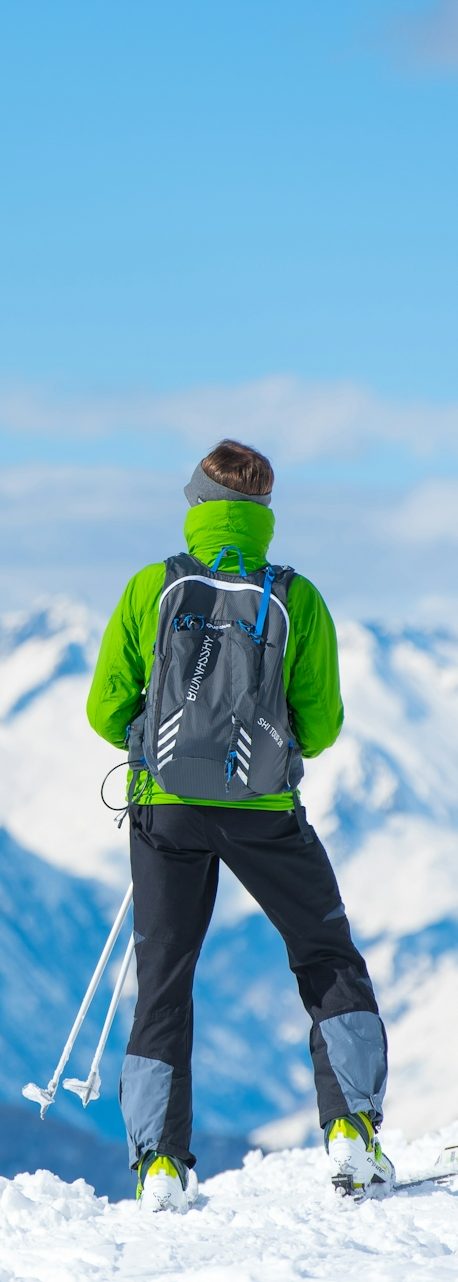Wanderer in grüner Jacke mit Rucksack blickt auf schneebedeckte Berge.